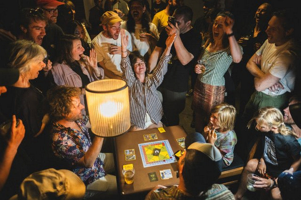 A woman celebrates triumphantly -  arms raised -  at a lively board game gathering with a joyful -  cheering crowd