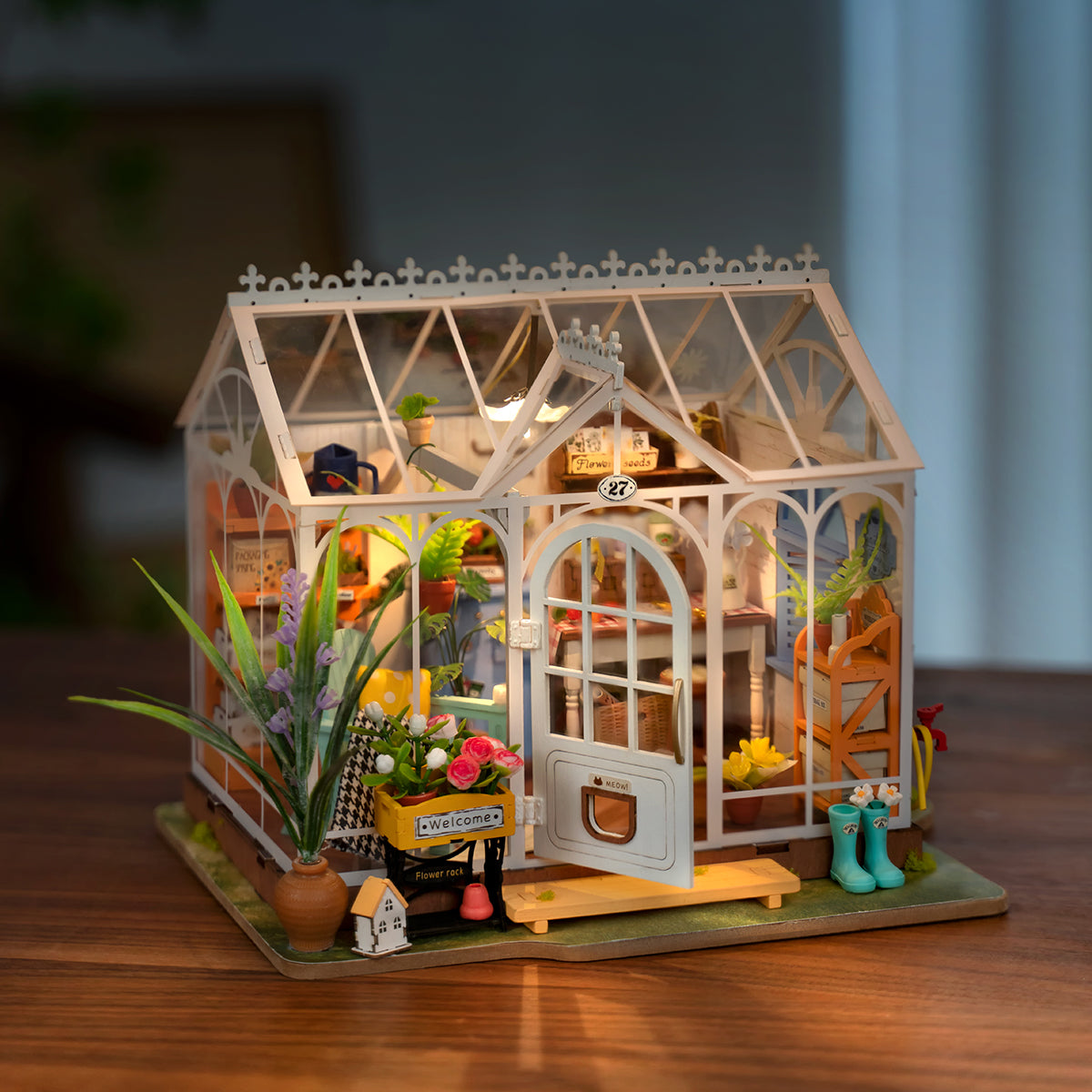 Illuminated miniature white greenhouse model filled with tiny plants, flowers, and gardening supplies