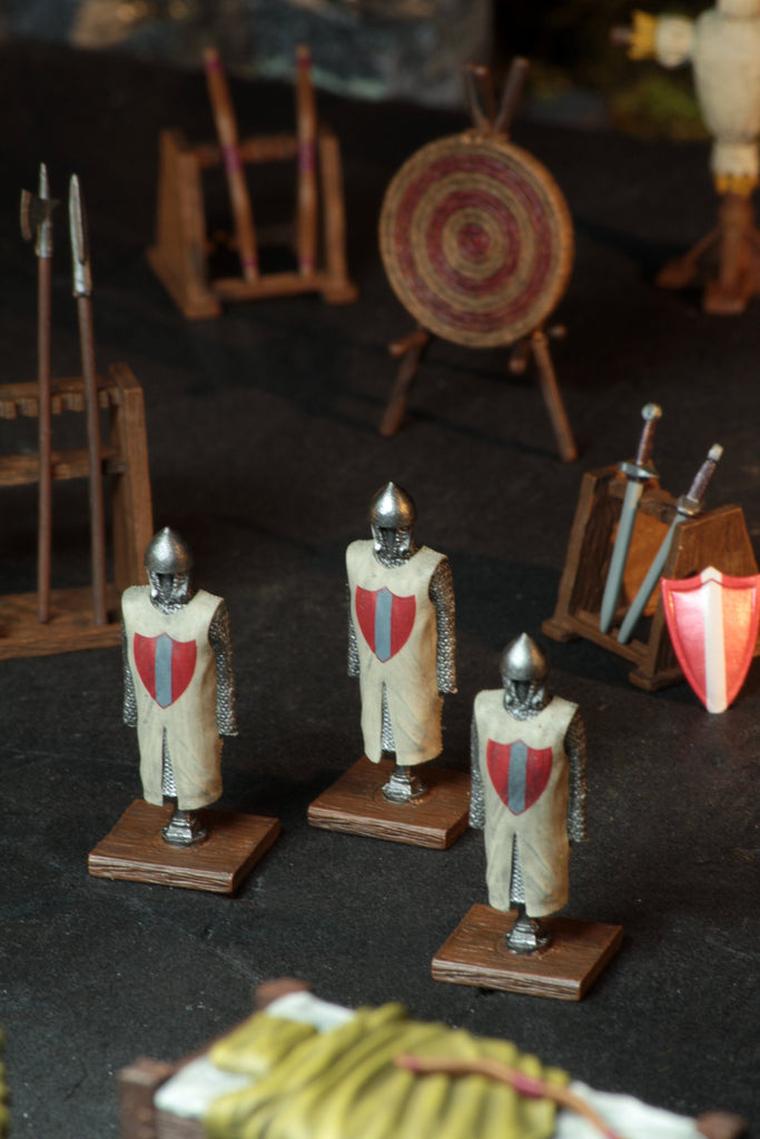 Three miniature medieval knights in chainmail and surcoats, surrounded by tiny archery target, spears, and swords