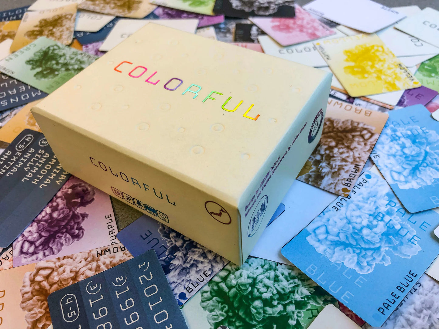 The colourful Card Game box with rainbow text rests on a collection of multiColoured cards featuring abstract textures and colour names