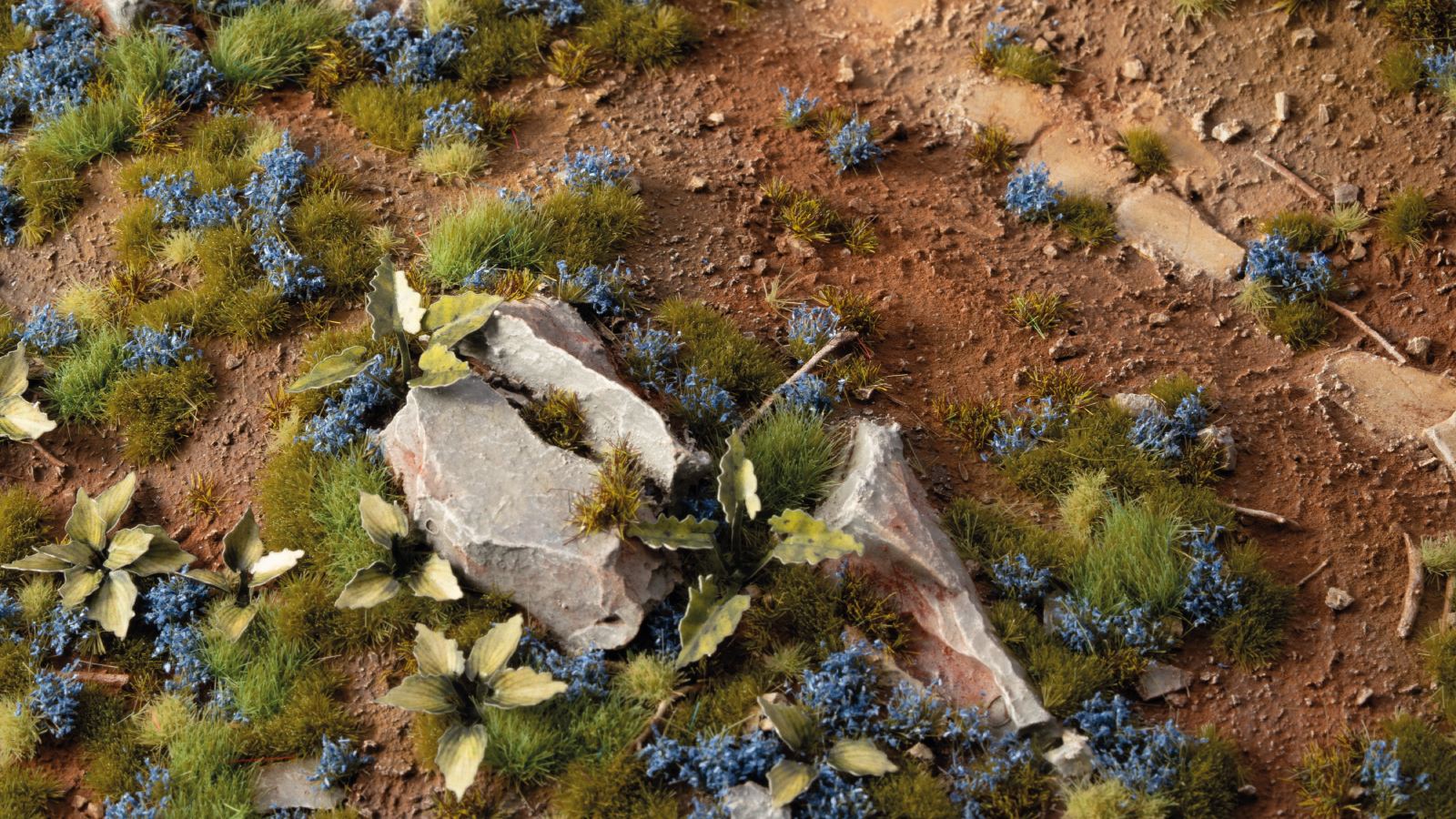 Miniature terrain diorama featuring reddish dirt, green grass, blue flowers, and jagged gray rocks