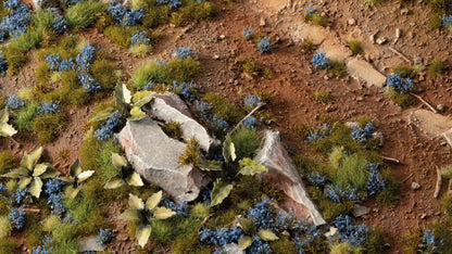 Miniature terrain diorama featuring reddish dirt, green grass, blue flowers, and jagged gray rocks