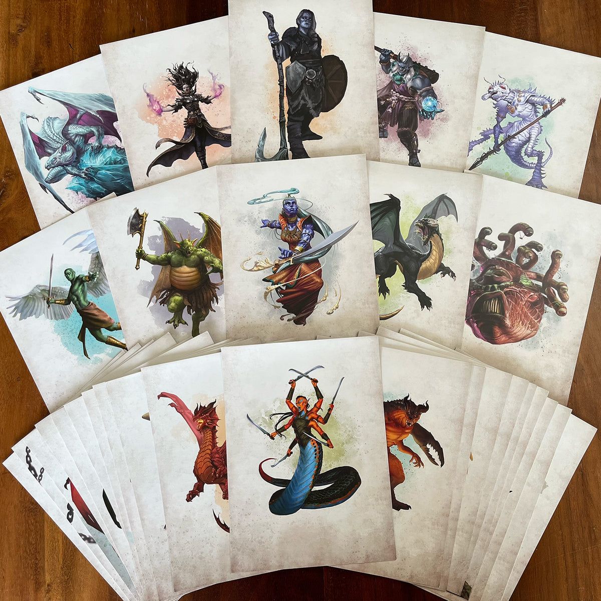 A fanned-out collection of fantasy art prints on a wooden table, featuring diverse characters and creatures: dragons, humanoids, multi-limbed beings, and monstrous figures