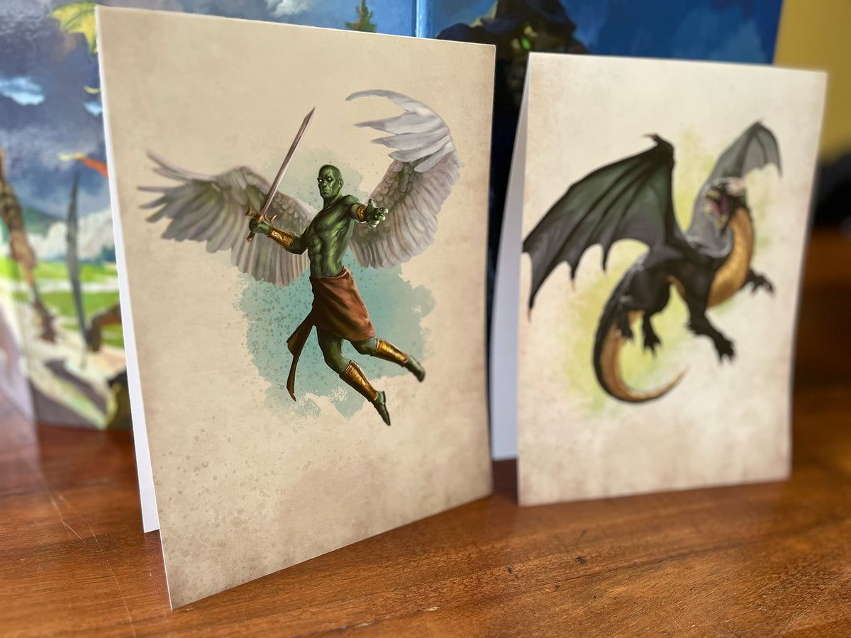 Two fantasy art prints on a wooden table: a green-skinned winged warrior with a sword and a black and gold dragon