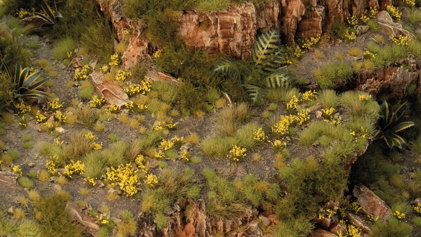 Miniature rocky landscape diorama with dense green grass, agave plants, and scattered bright yellow flowers