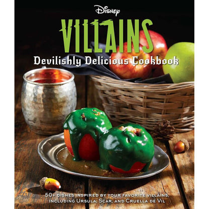 The Disney Villains Devilishly Delicious Cookbook cover displays two red apples coated in green icing on a plate beside a silver mug and a basket of apples