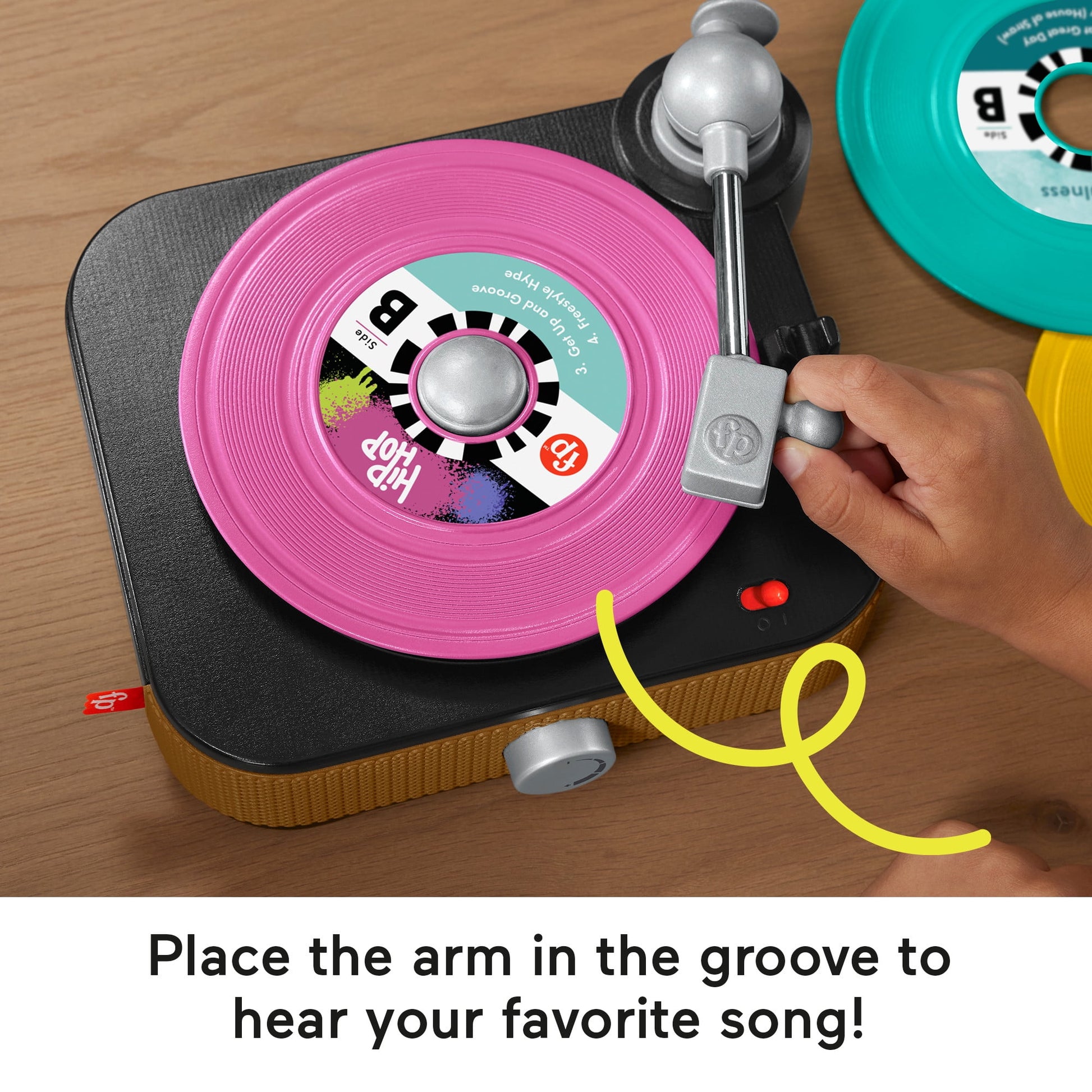 A child's hand lowers the arm onto a bright pink record on a toy turntable. Other colorful records are nearby