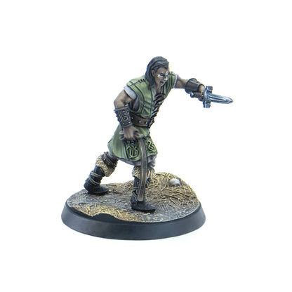 A grimacing male warrior miniature in green and leather armor lunges, wielding a dagger forward and a curved blade