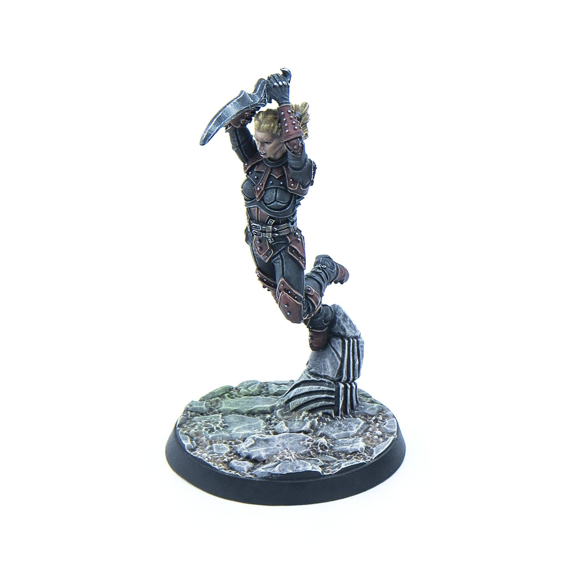 A blonde female warrior miniature in dark armor, leaping from a rocky base with a curved blade raised to strike