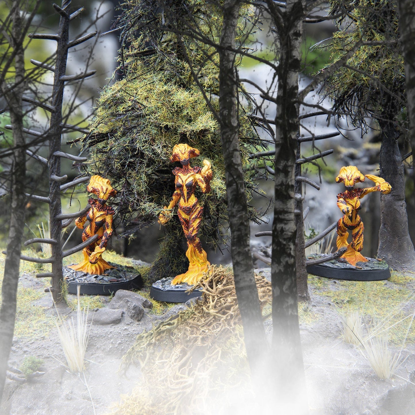 Three vibrant orange and yellow flame-like miniatures stand in a misty, moss-covered miniature forest
