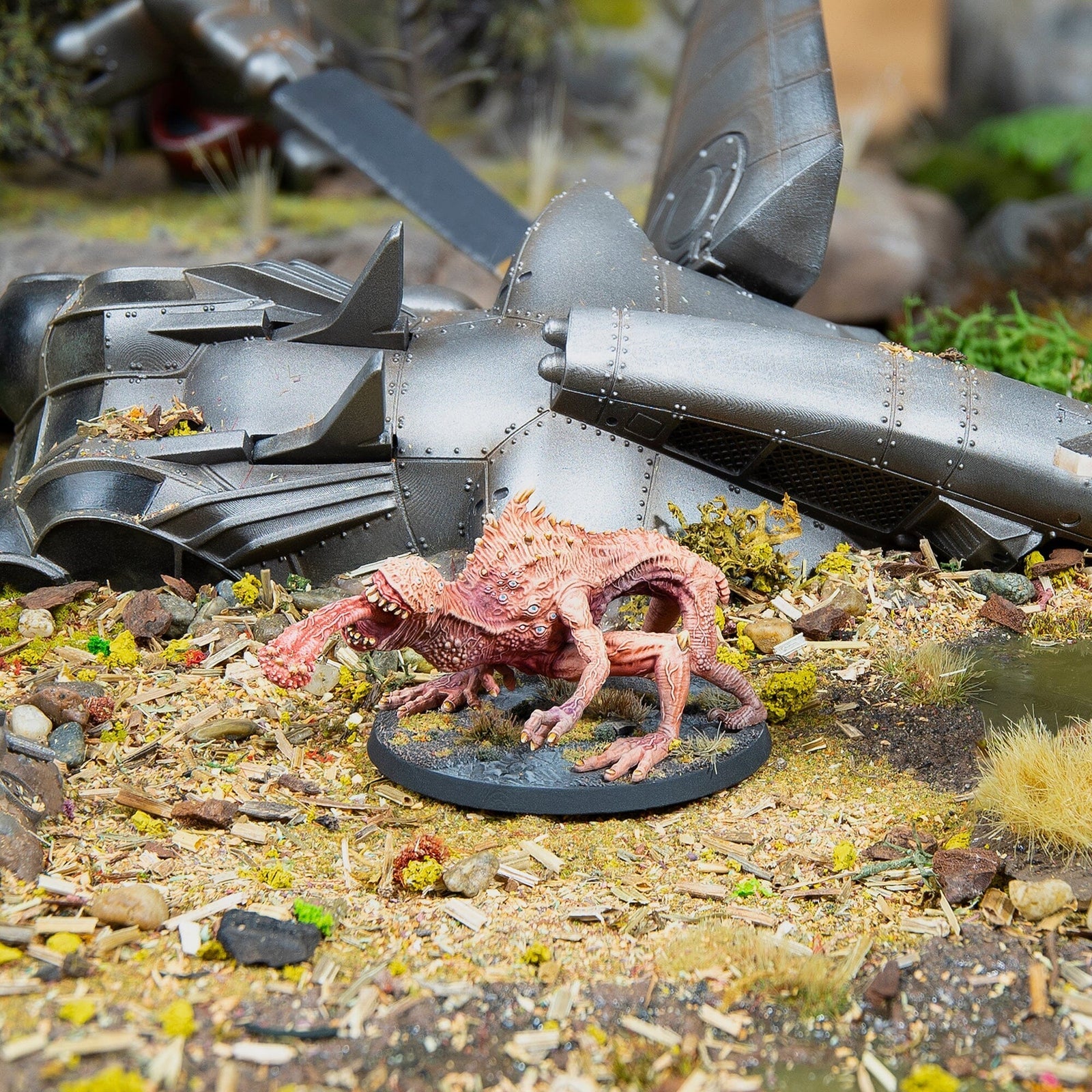Fleshy, pink monster miniature stands on a diorama base next to large metallic wreckage
