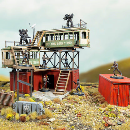 Miniature wargame scene: Armed figures defend a wasteland fort made of red containers and a salvaged tram car