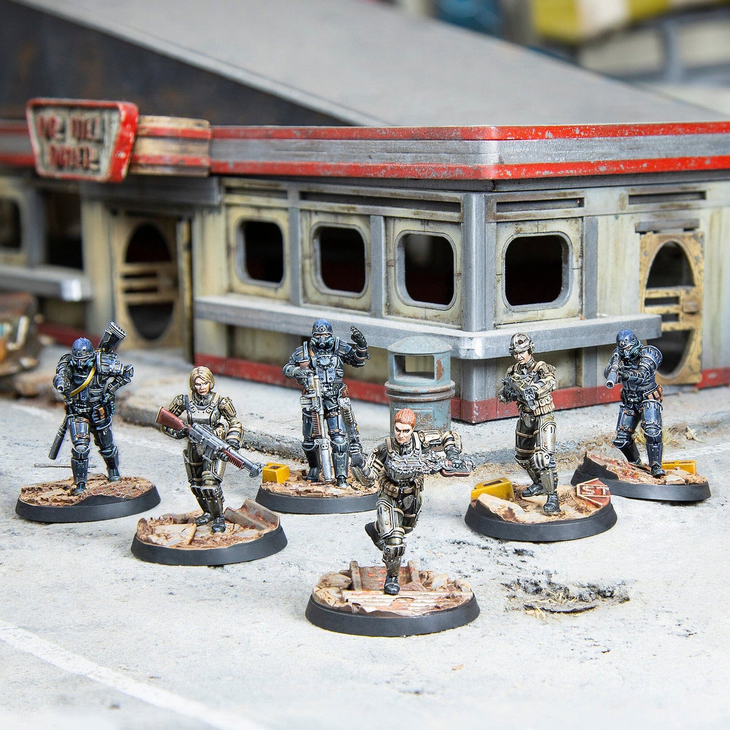 Six miniature figures from Fallout Wasteland Warfare - Brotherhood of Steel Combat Patrol stand on a concrete surface in front of a weathered building