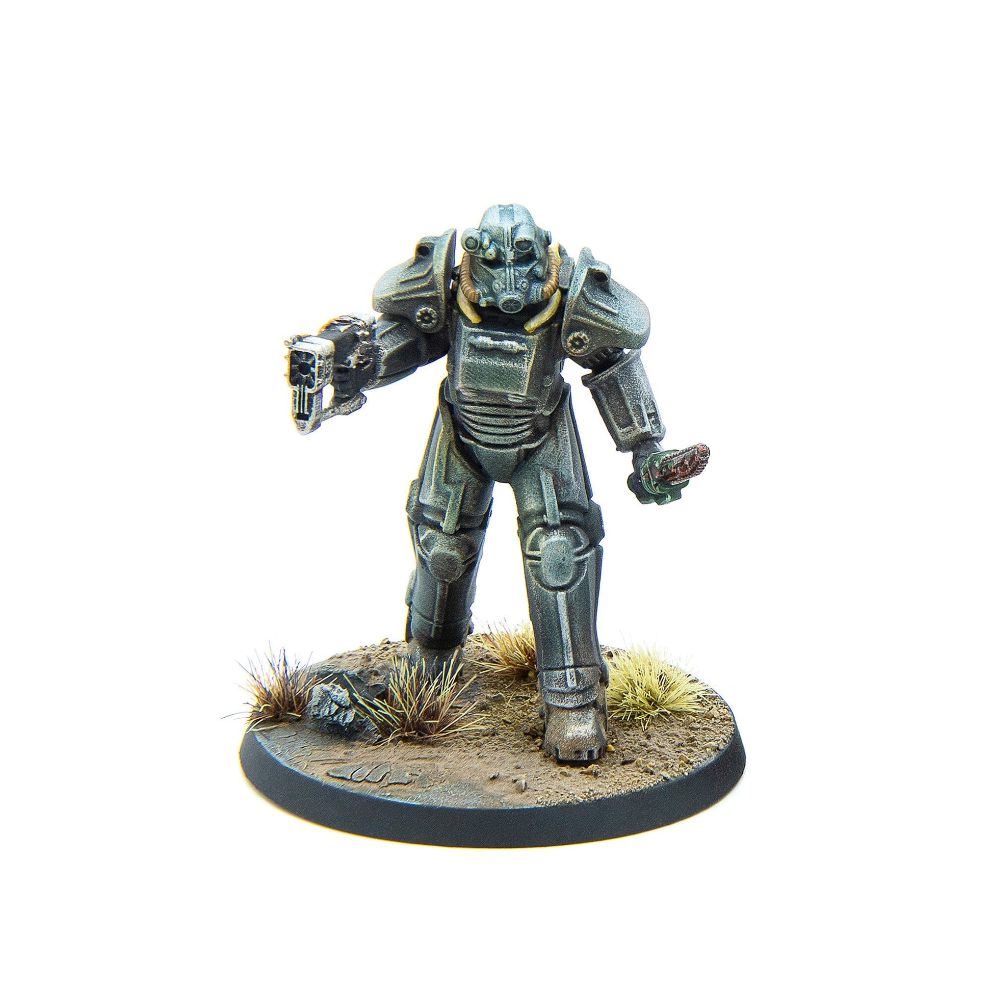 A tabletop miniature of a grey Fallout power armor figure holding a gun and an object, standing on a dirt base with dry grass