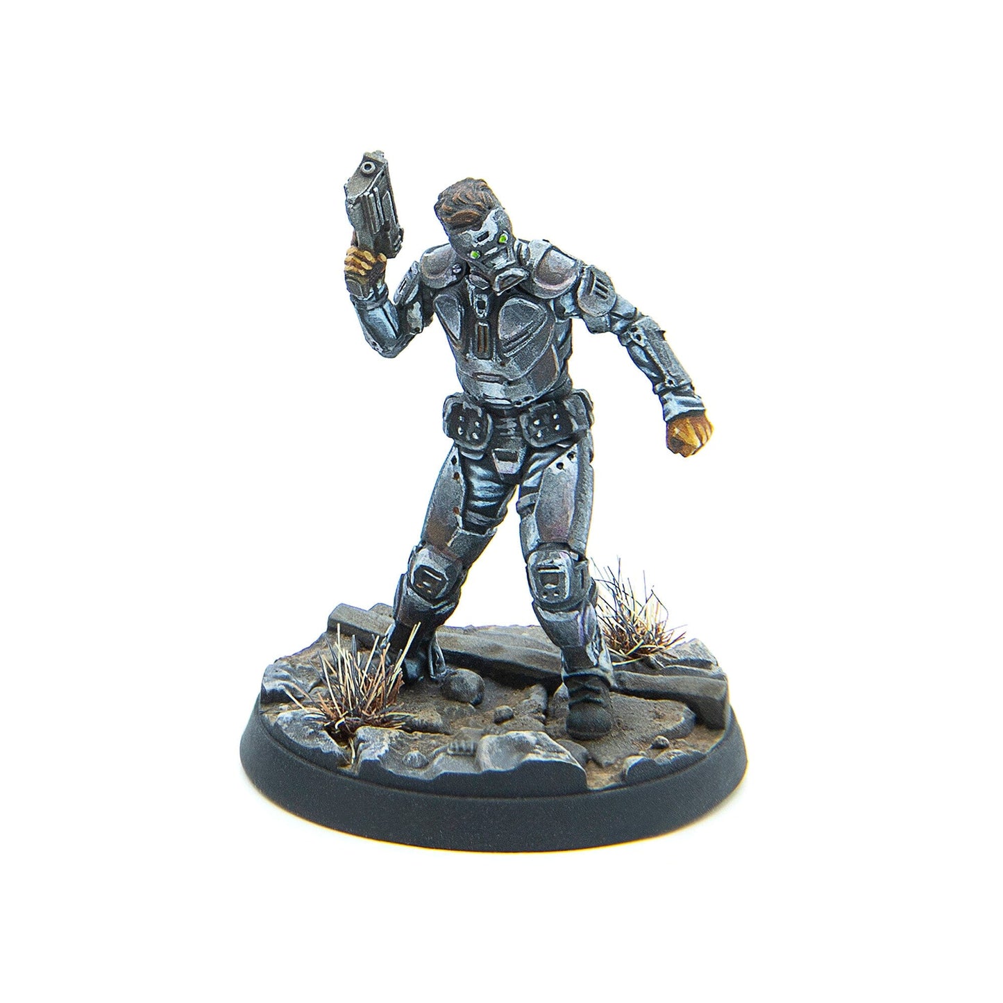 A silver-armored sci-fi miniature with green glowing eyes holds a pistol up, standing on a rocky, grassy base