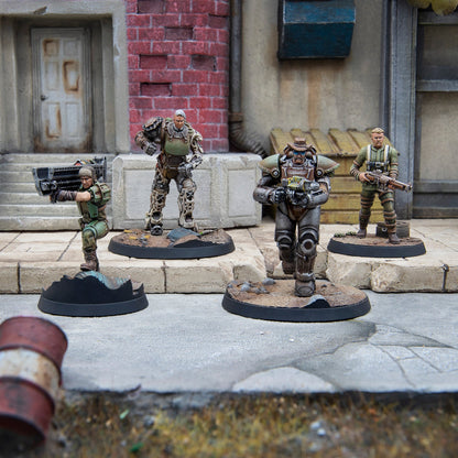 Four Fallout wargaming miniatures in an urban diorama: a soldier aims a heavy weapon, two wear power armor, one holds a rifle