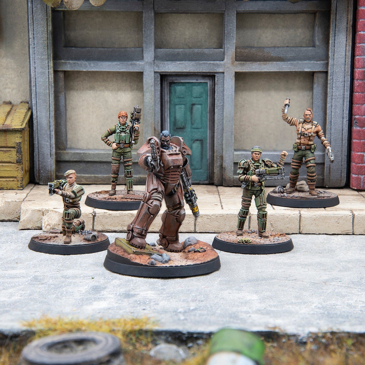 Five detailed post-apocalyptic miniatures, led by a power-armored figure, stand armed in a ruined city