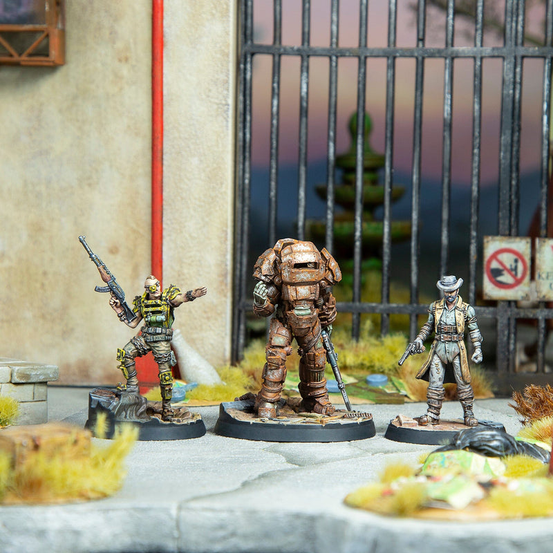Three tabletop miniatures: a raider with a rifle, rusty power armor, and a cowboy with pistols, in a wasteland scene