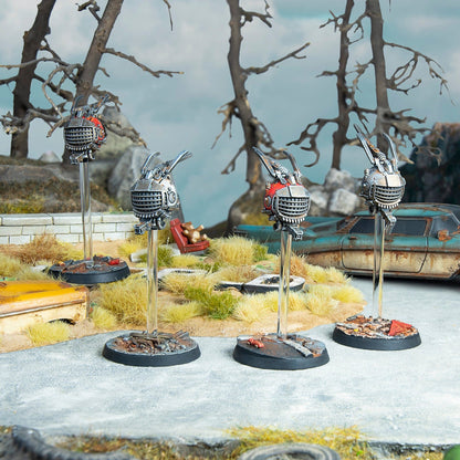 Four miniature metallic robots, three silver and one red, hover over a post-apocalyptic scene with dead trees and a rusty car