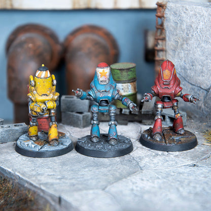 Three weathered robot miniatures: yellow, blue, and red, stand on bases in an industrial environment