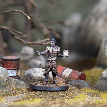A painted miniature of a dark-skinned person in ragged clothes, holding a vial and a clipboard, in a wasteland with rusted barrels
