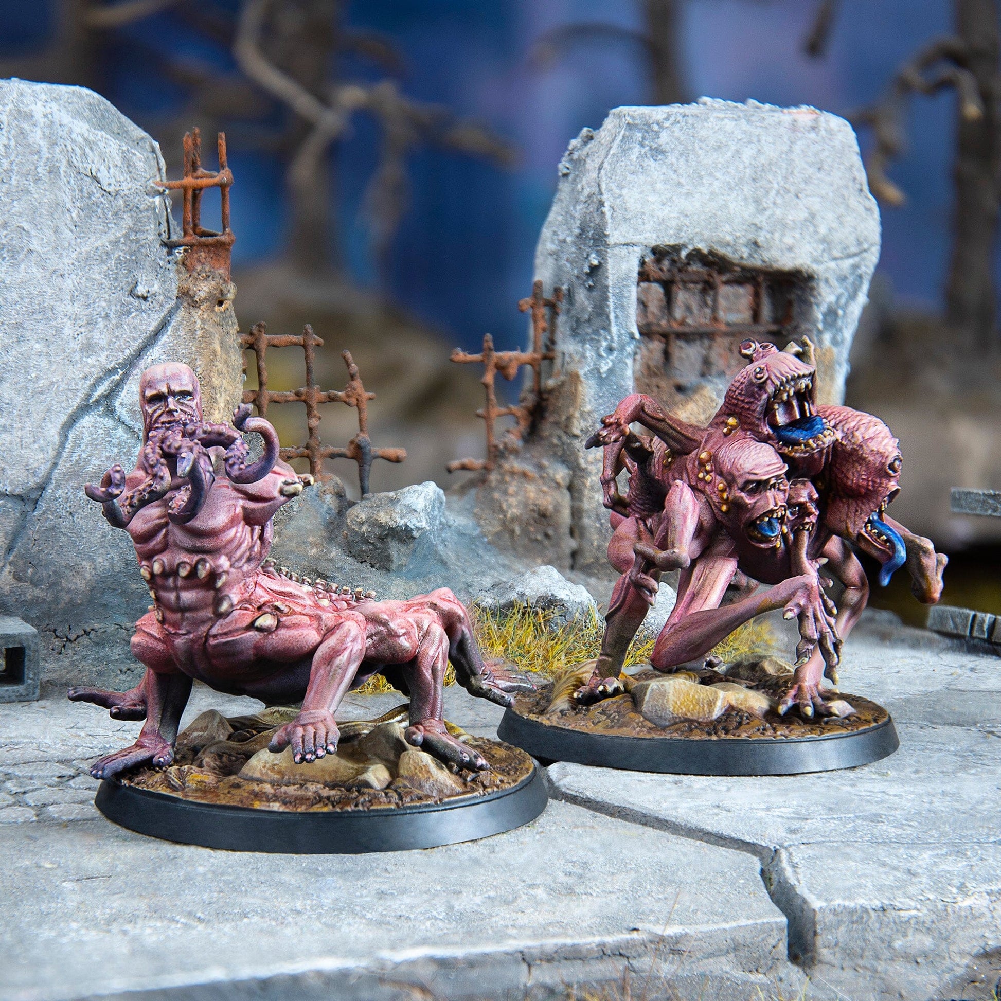 Two painted Fallout Wasteland Warfare Super Mutants Centaurs miniatures, one with tentacles around its head and the other with multiple heads, stand on a concrete base with ruins in the background