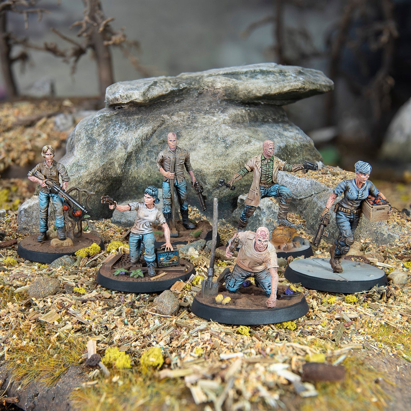 Six post-apocalyptic miniature figures, some with ghoul-like faces, are shown scavenging in a rocky wasteland