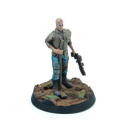 A miniature of a mutated man in worn clothes, holding a staff and a gun, on an earthy base with scattered corn
