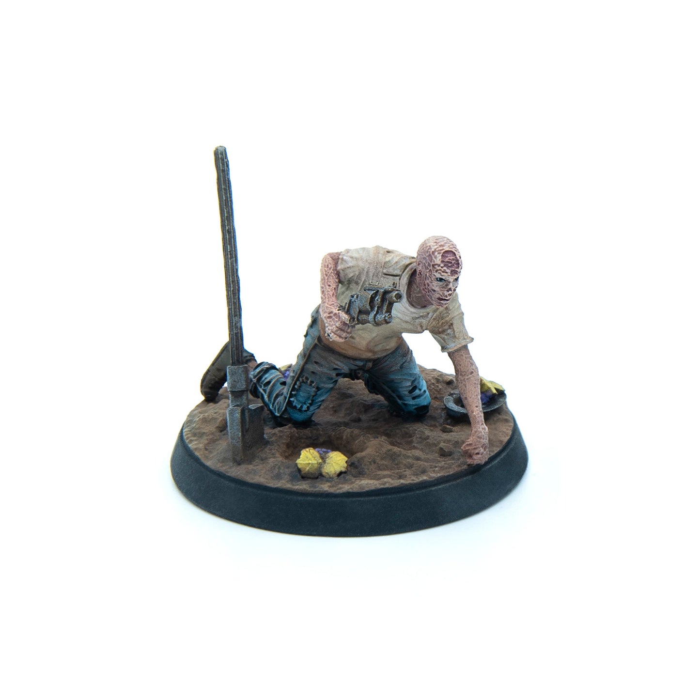 A severely scarred miniature man crouches, holding a crude weapon, on a sandy terrain base with a shovel