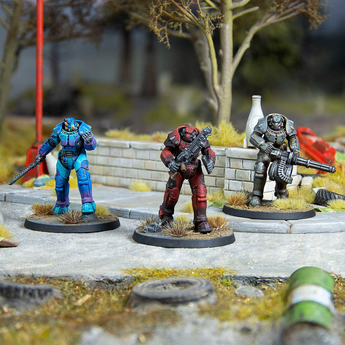 Three power-armored soldier miniatures: blue with blade, red with rifle, grey with minigun, on an urban diorama