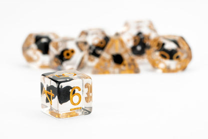 Clear resin D6 die with black and gold swirls, showing the number 6, in front of a blurred set of polyhedral dice
