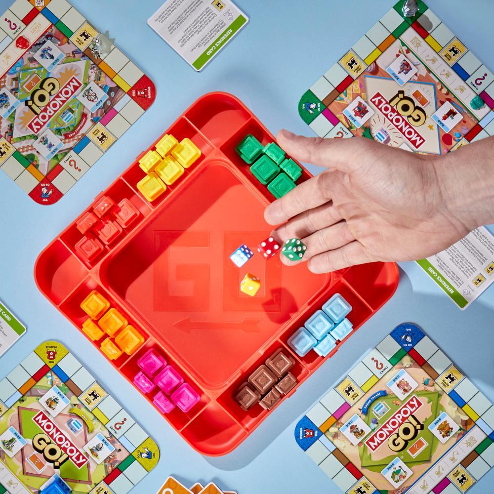 A hand rolls dice into a red game piece tray surrounded by Monopoly GO! game boards and various colorful game pieces