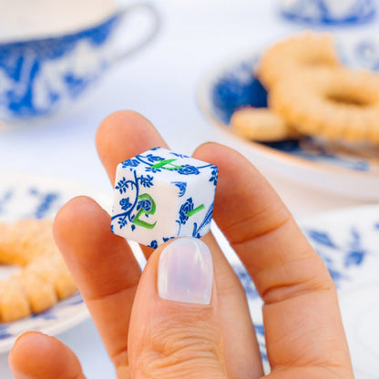 A hand holds a white RPG dice with blue floral patterns and green numbers, visible against a blurred background of blue and white tea party dishes and cookies
