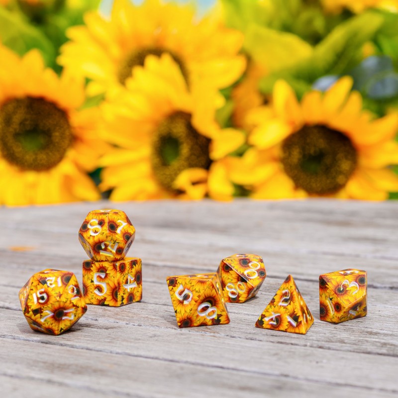 A Full Art Sunflower RPG Dice Set, yellow with sunflower patterns and white numbers, on a wooden surface with blurred sunflowers in the background