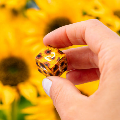 A hand holds a sunflower-patterned RPG die, showing a 2 on top, against a blurred background of yellow sunflowers