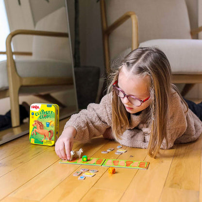 A young girl with glasses lies on a wooden floor, playing the HABA Clippety-Clop! board game with a horse figurine.