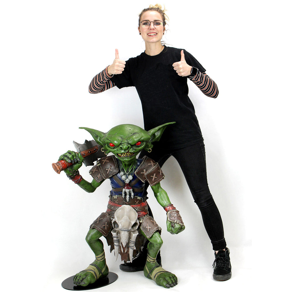 A smiling woman gives two thumbs up next to a large, green, axe-wielding goblin statue