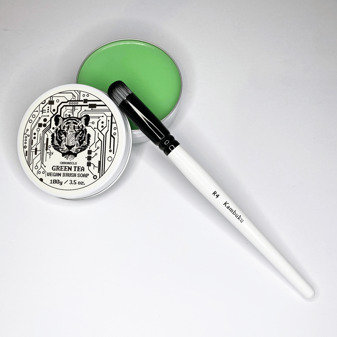 Set of makeup brush cleaners: open tin of green vegan soap, its lid with a cybernetic tiger design, and a black textured silicone mat