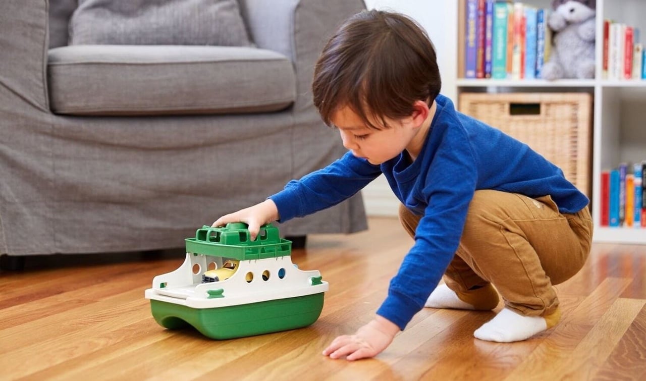 Boy in blue shirt squatting on a wooden floor, playing intently with a green and white toy ferry boat