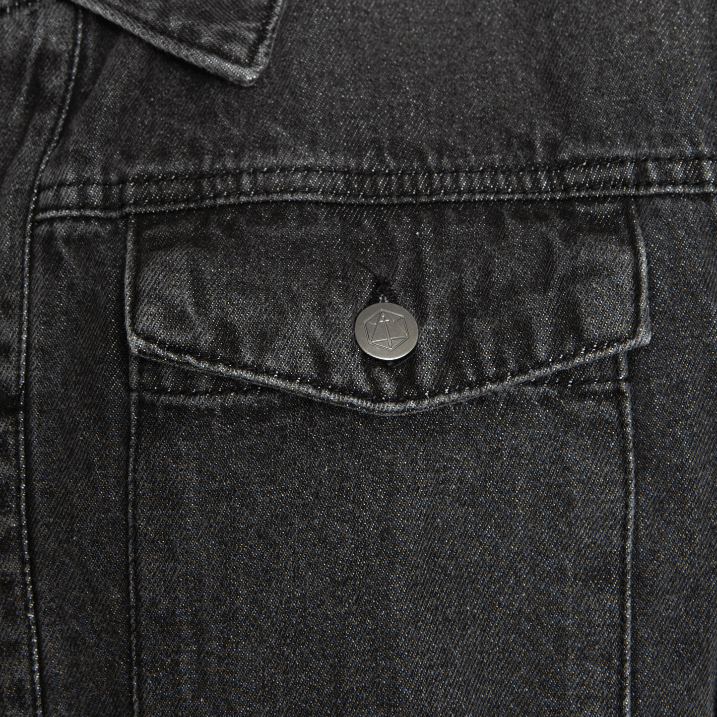 Dark grey denim jacket pocket with a metallic button showing a sword in a hexagon