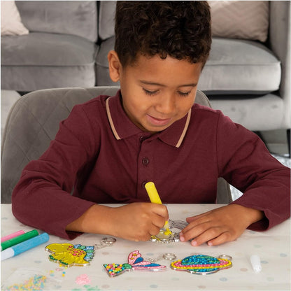 Smiling boy colouring a plastic keychain design with a yellow marker. Colourful finished crafts are on the table