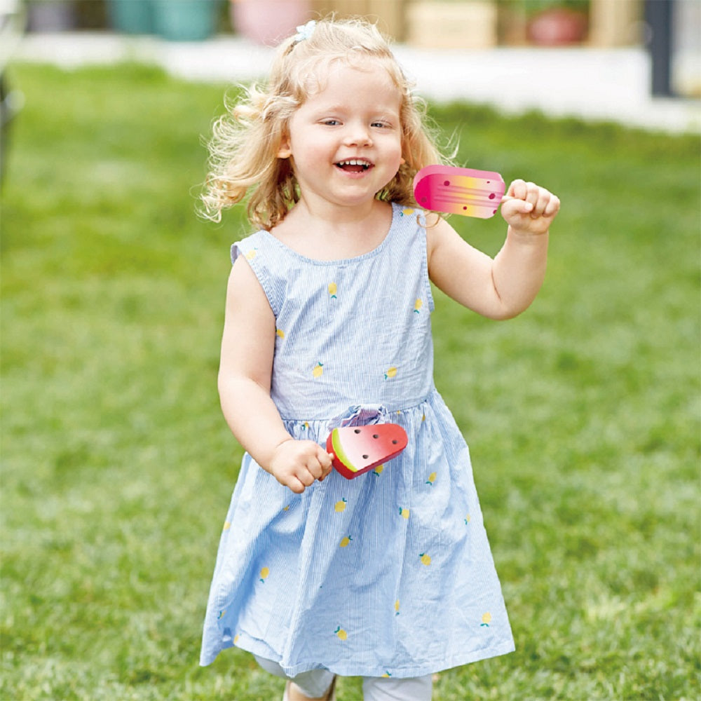 Smiling blonde toddler holding two wooden toy popsicles (watermelon and pink) outside on grass