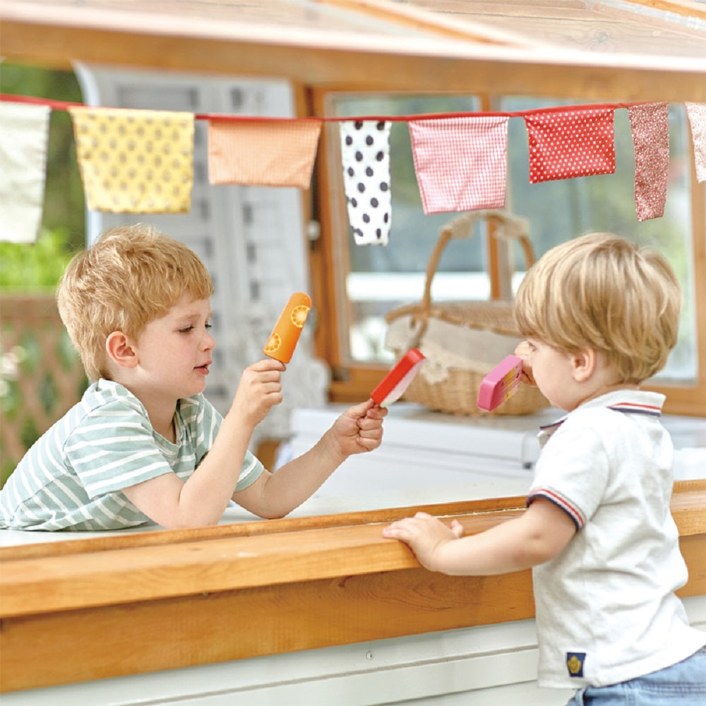 Two boys play with toy popsicles across a wooden stand decorated with fabric bunting