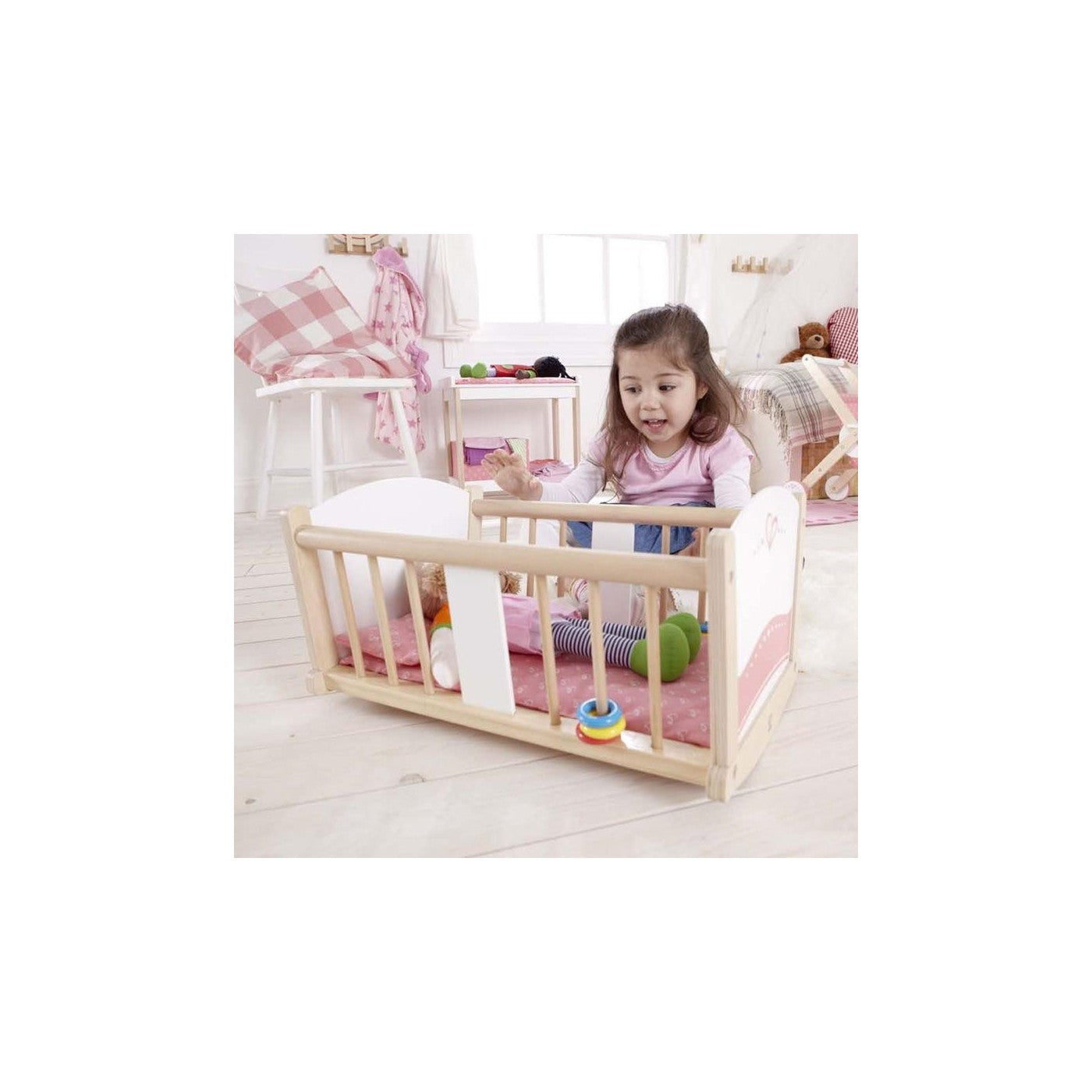 A young girl smiles while leaning over a wooden doll's crib with a doll and pink bedding.