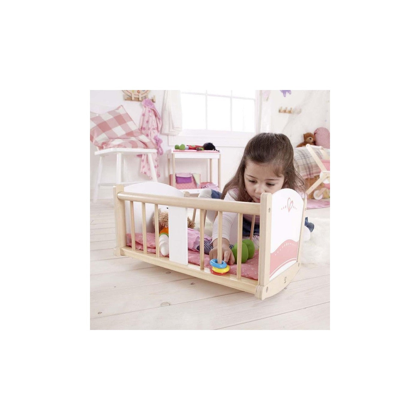 A young girl plays with a colorful stacking ring inside a wooden doll's crib, which also holds a doll.