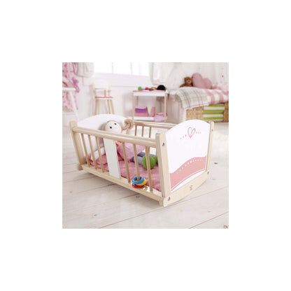 Wooden toy rocking cradle with white and pink ends. A rag doll lies on a pink mattress inside, with a small colorful toy.