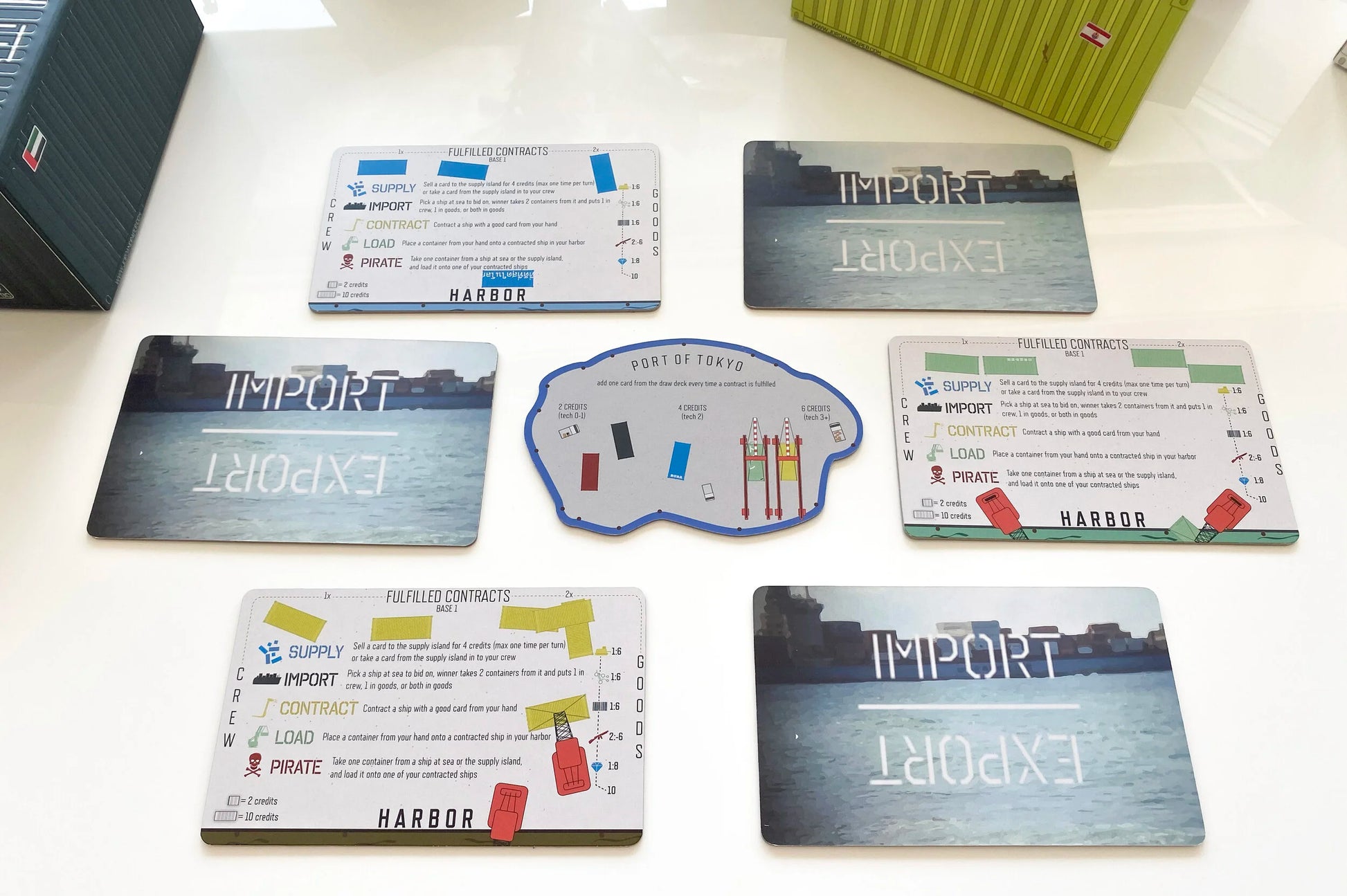 A collection of Import_Export and Harbor board game cards, along with a Port of Tokyo game board, arranged on a white surface with game boxes in the background