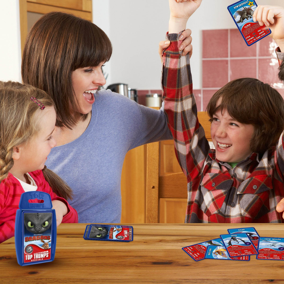 A boy triumphantly raises a Dragon Top Trumps card while his mother cheers and sister watches at a wooden table