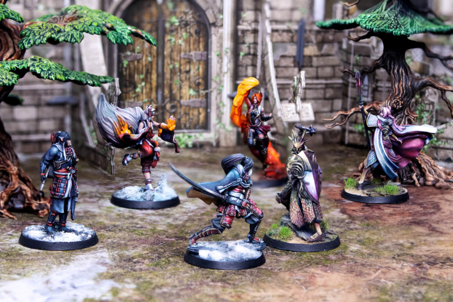 Six painted fantasy miniatures in dynamic poses on a tabletop terrain with trees and ruins