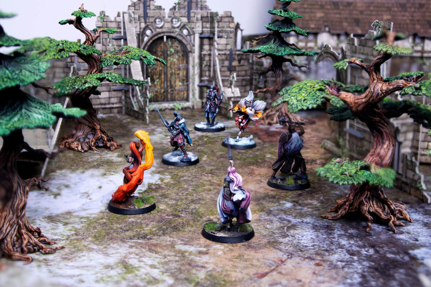 Fantasy miniatures (warriors, mages) arranged on a tabletop game mat with trees and castle ruins